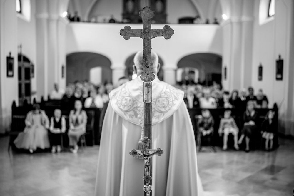 Black and white view from behind a priest holding a crucifix during a church service with congregation in background.