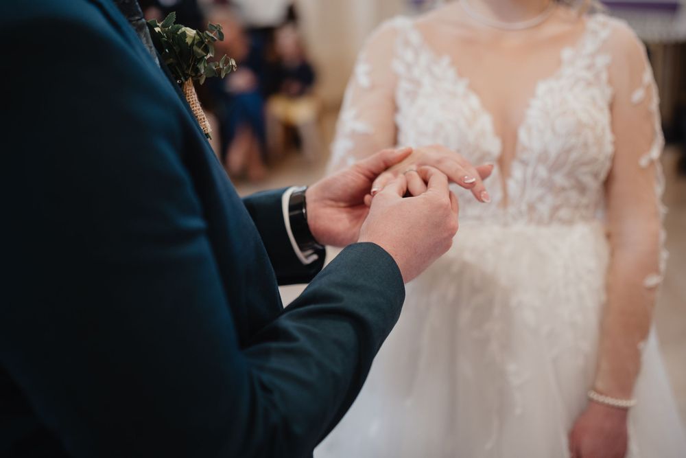 Close-up of groom holding wedding ring during ceremony with bride in lace and tulle dress in background.