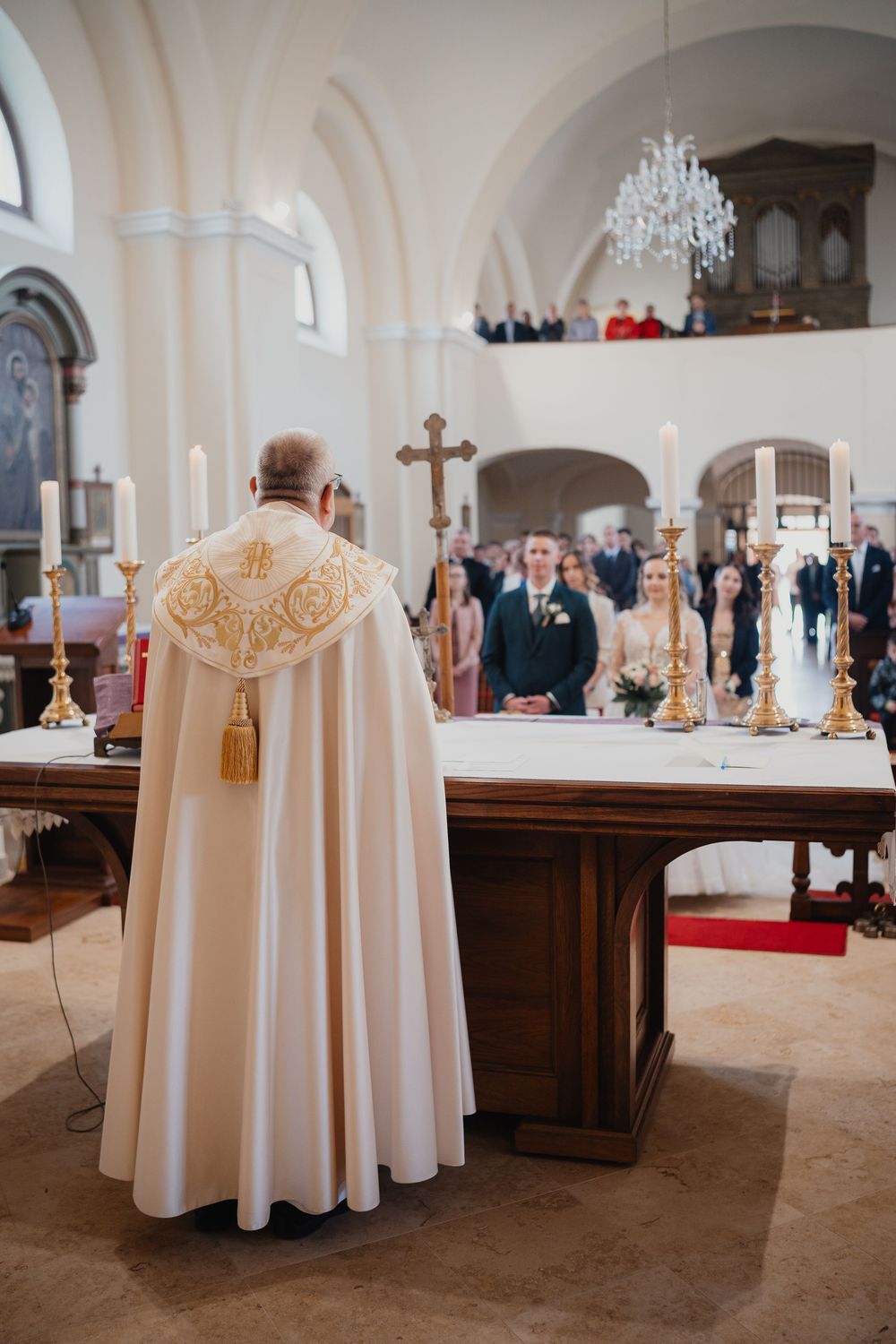 Catholic priest in ornate cream vestments leads traditional church ceremony at altar with crystal chandelier above.