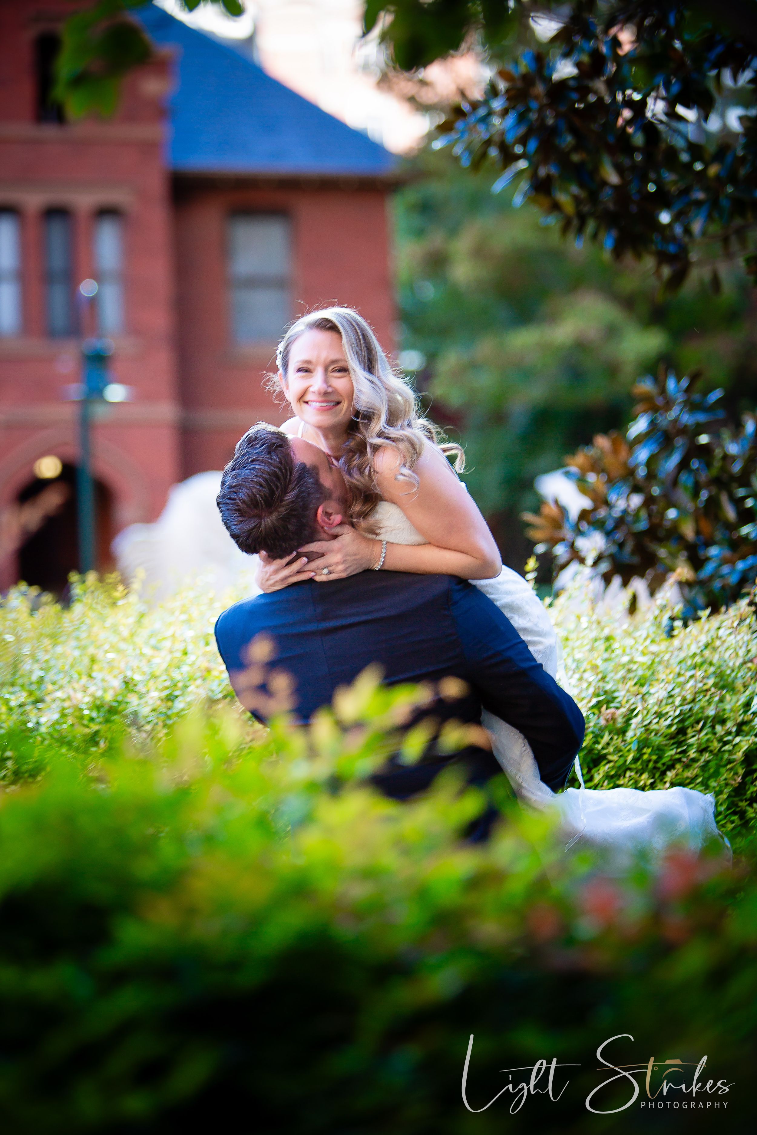 A joyful couple embraces in a garden with a brick building in the background during an outdoor engagement photo session.