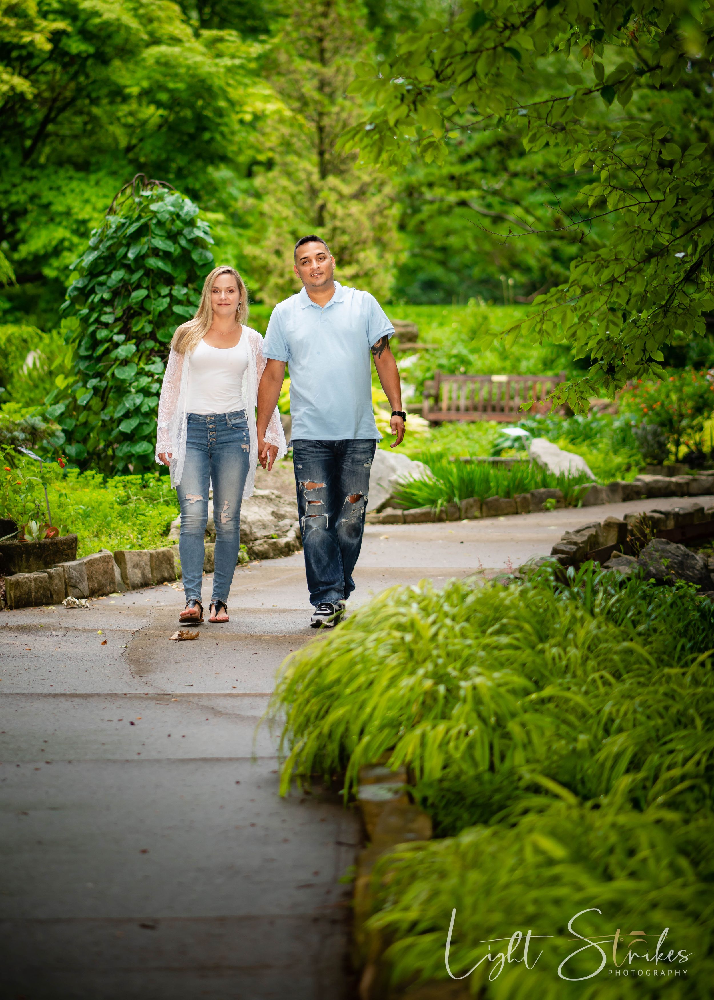 Engagement session of a couple walking hand in hand along a winding garden path surrounded by lush green foliage and landscaping.