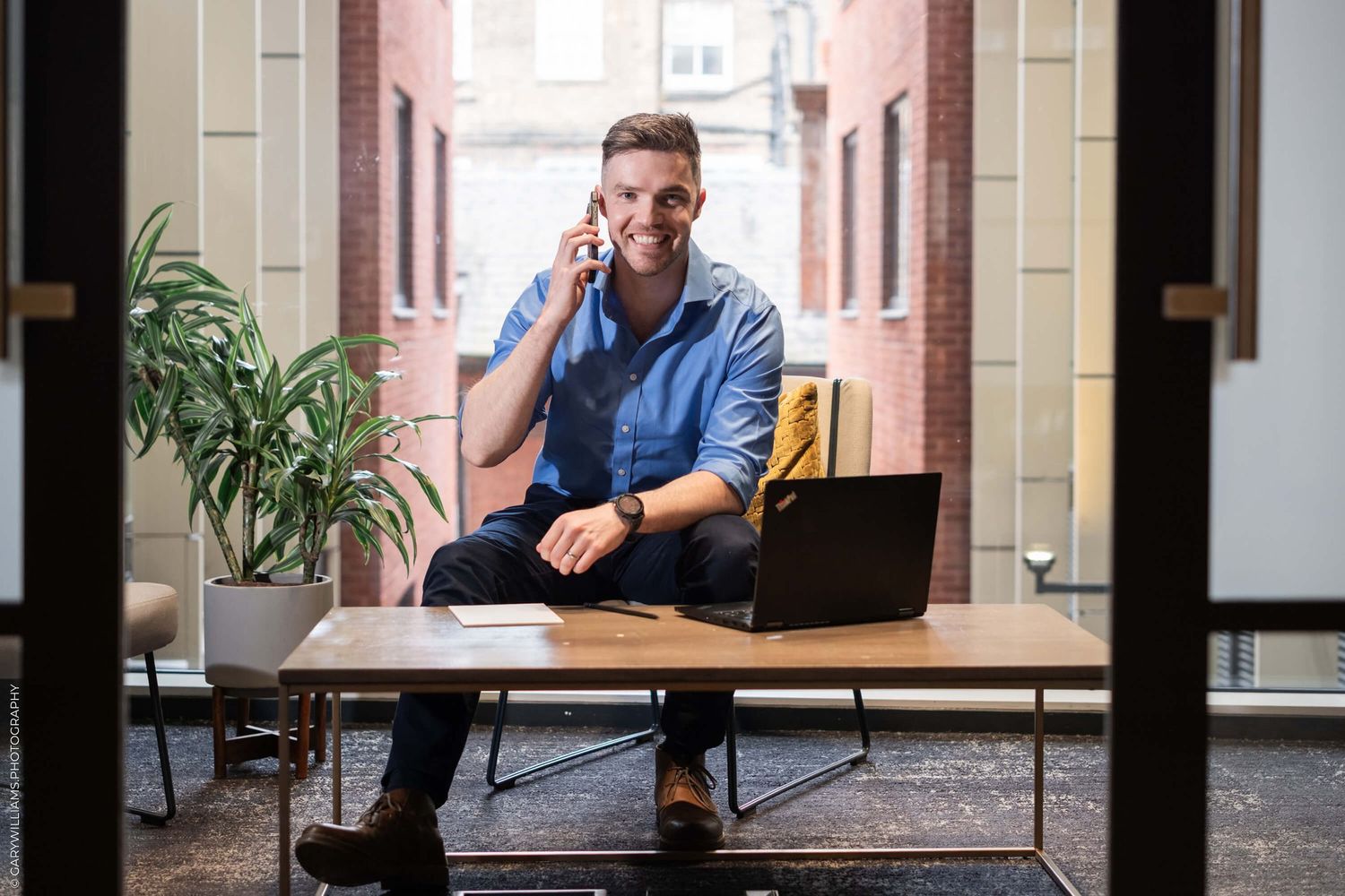 Professional in a blue shirt sits at desk in hallway with laptop while talking on mobile phone in office setting.