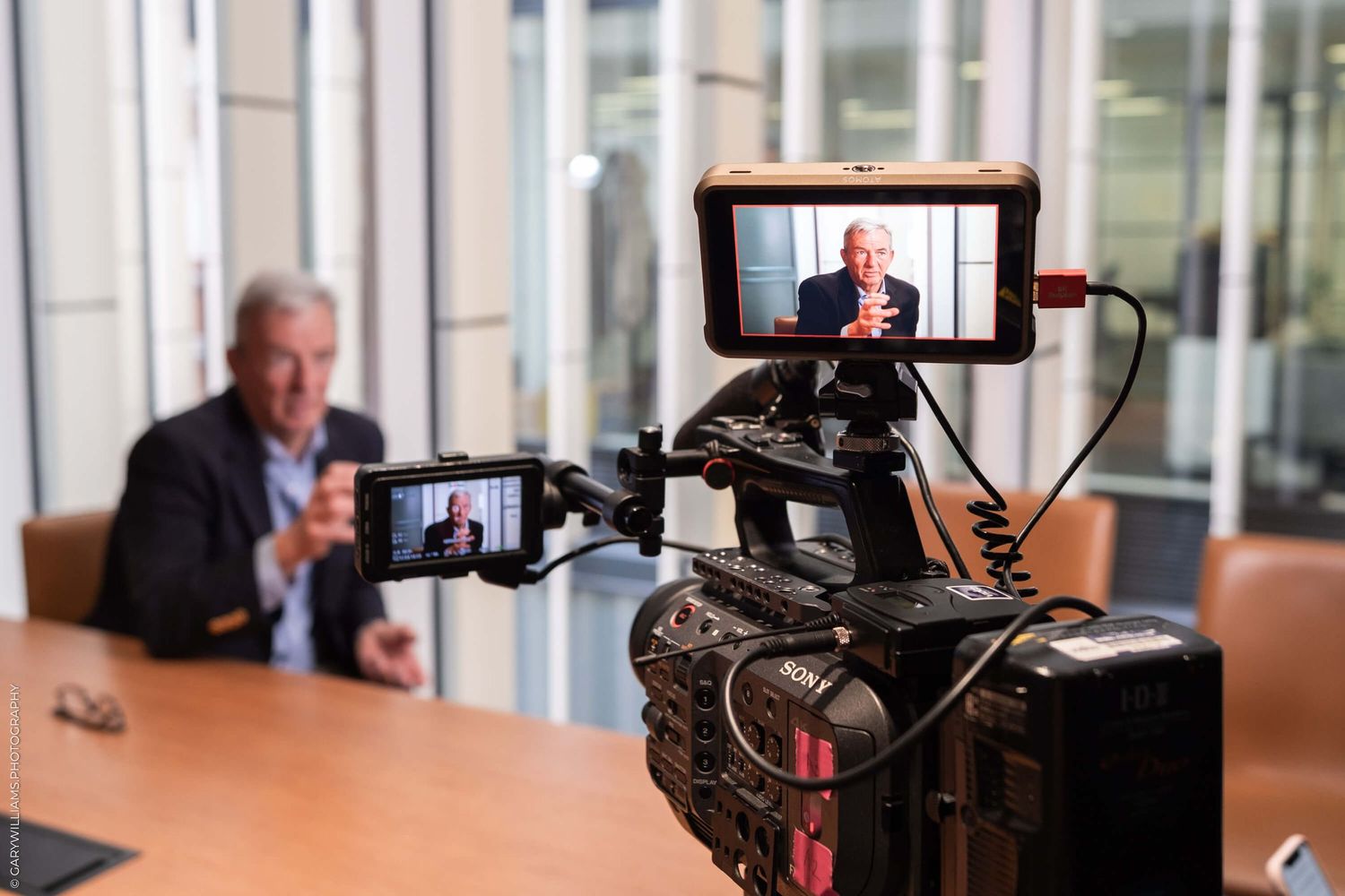 Professional video camera setup filming an interview at a conference table with dual LCD monitor displays.