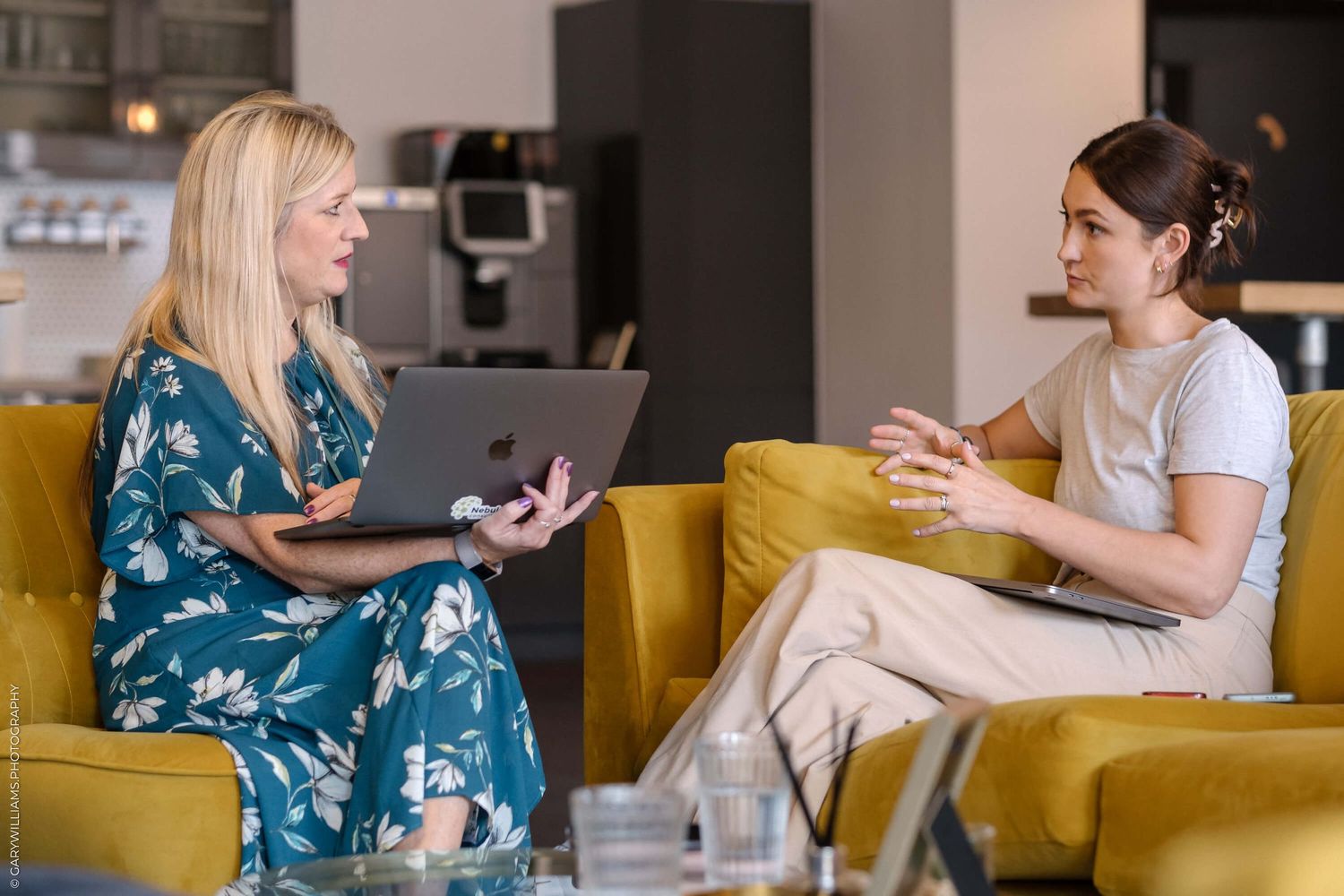Two people having a conversation while sitting on yellow couch with laptops.