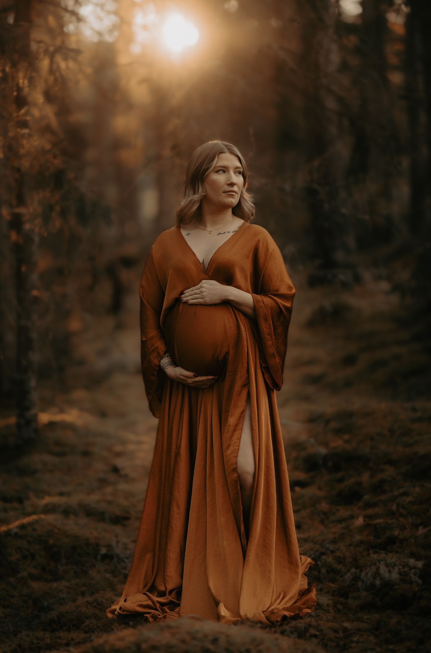 Maternity portrait of a woman in a flowing rust-colored dress standing in an autumn forest.