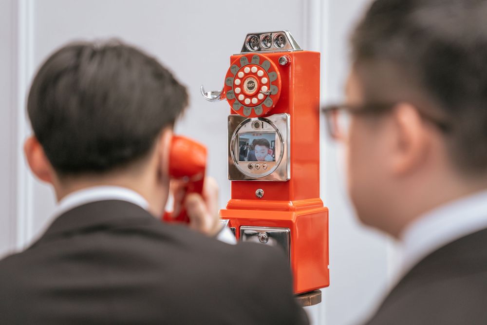 Two people in business attire examine a bright orange vintage rotary payphone mounted on a wall.
