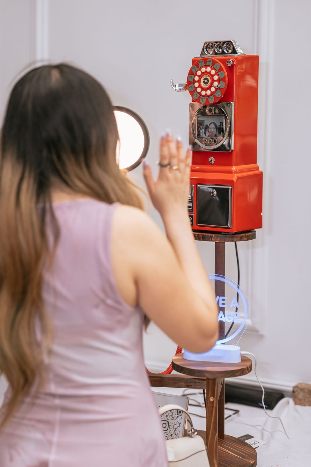 Woman in pink dress looks at vintage red rotary payphone mounted on wall in retro-styled interior setting.