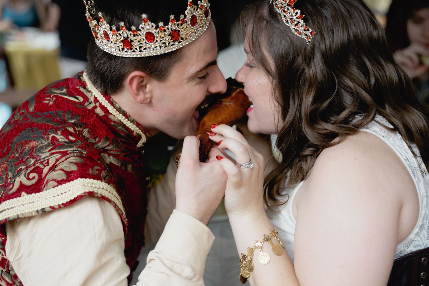 Two people in crown and formal attire sharing a bite of food during what appears to be a royal-themed celebration.