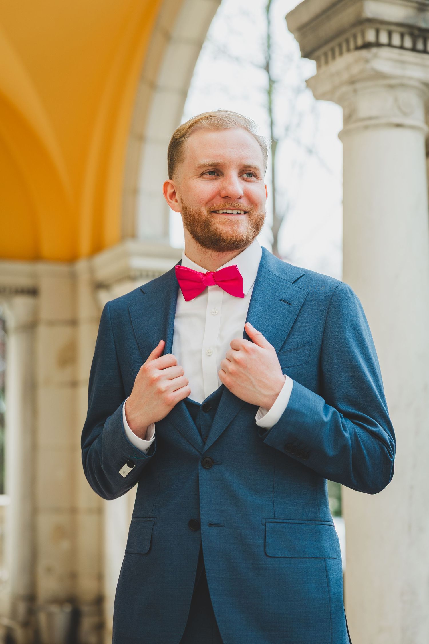 Groom in navy suit adjusts bright pink bow tie while standing against white pillared backdrop at formal event