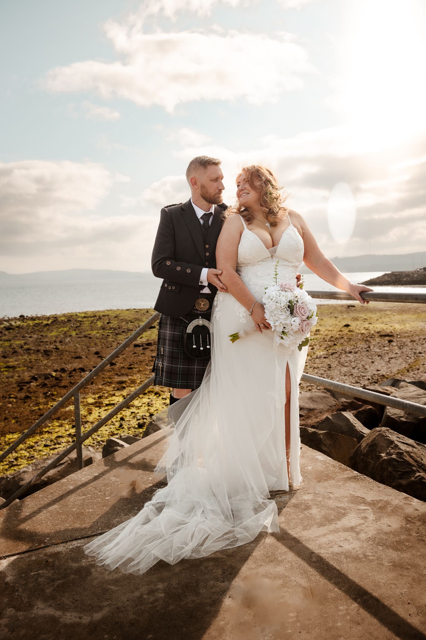 A couple in wedding attire standing on rocky coastal terrain with cloudy sky in the background.