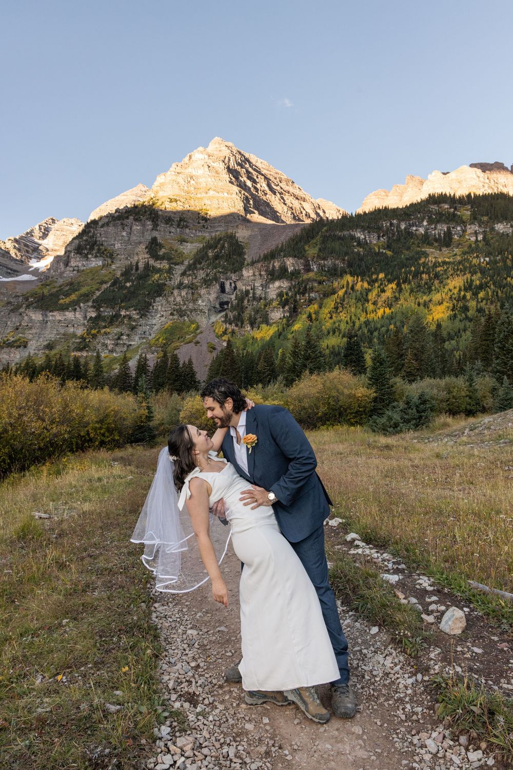 A couple in wedding attire share a romantic kiss along a mountain trail at sunset in the Rocky Mountains.