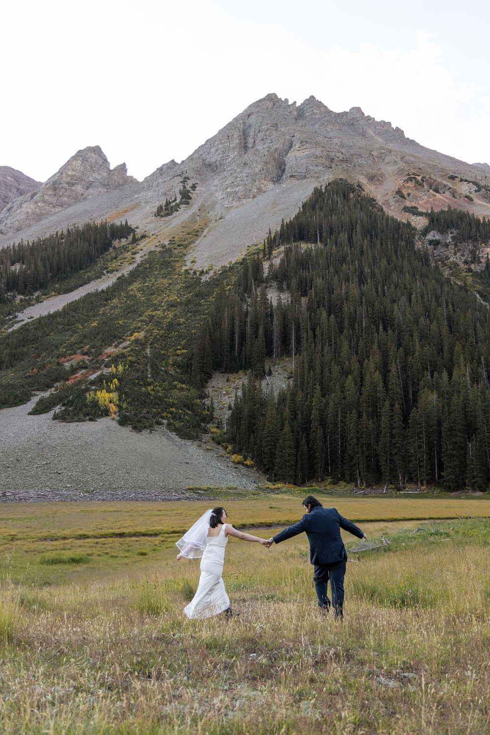 A couple in wedding attire runs through a mountain meadow with dramatic Rocky Mountain peaks rising in the background.