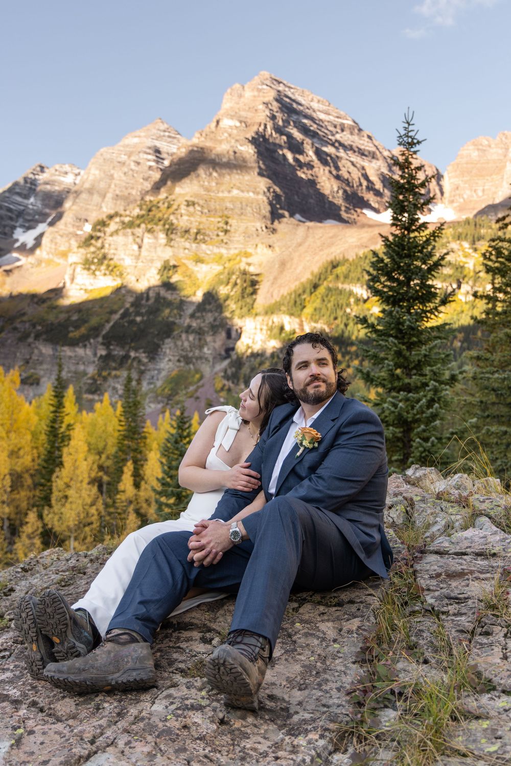 A couple in wedding attire sits on rocky terrain with majestic mountain peaks and autumn trees in the background.