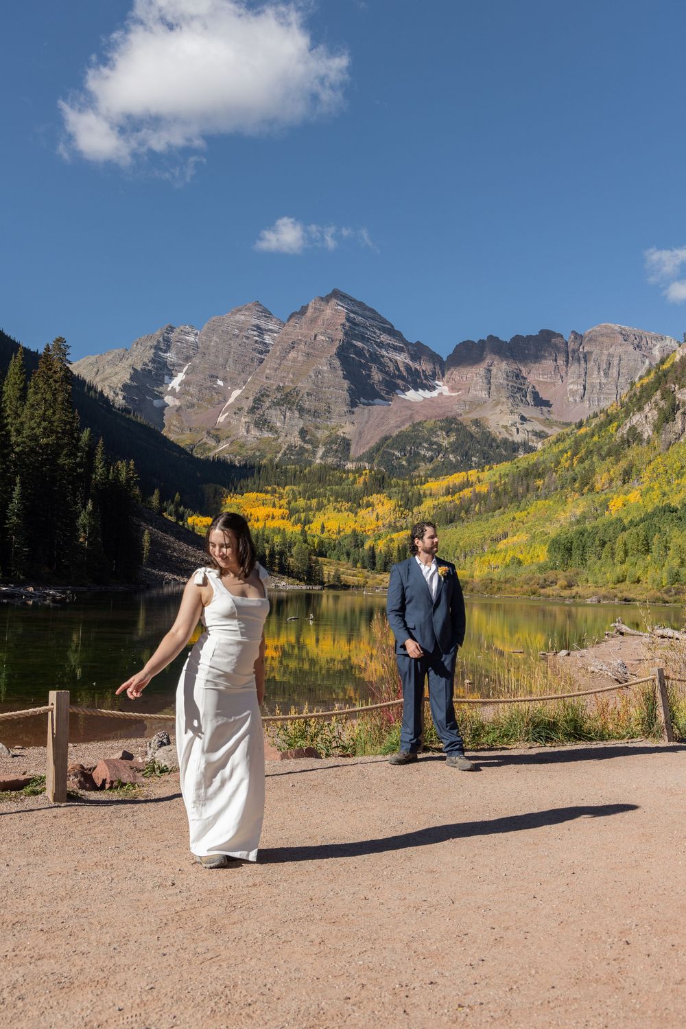 Couple in wedding attire standing at Maroon Bells lake in Colorado with fall foliage and mountain peaks in background.