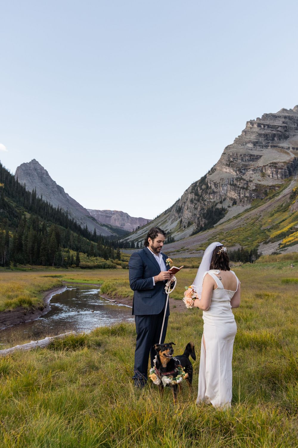 A couple celebrates their wedding day in a scenic mountain meadow with dramatic peaks and a winding stream in the background.