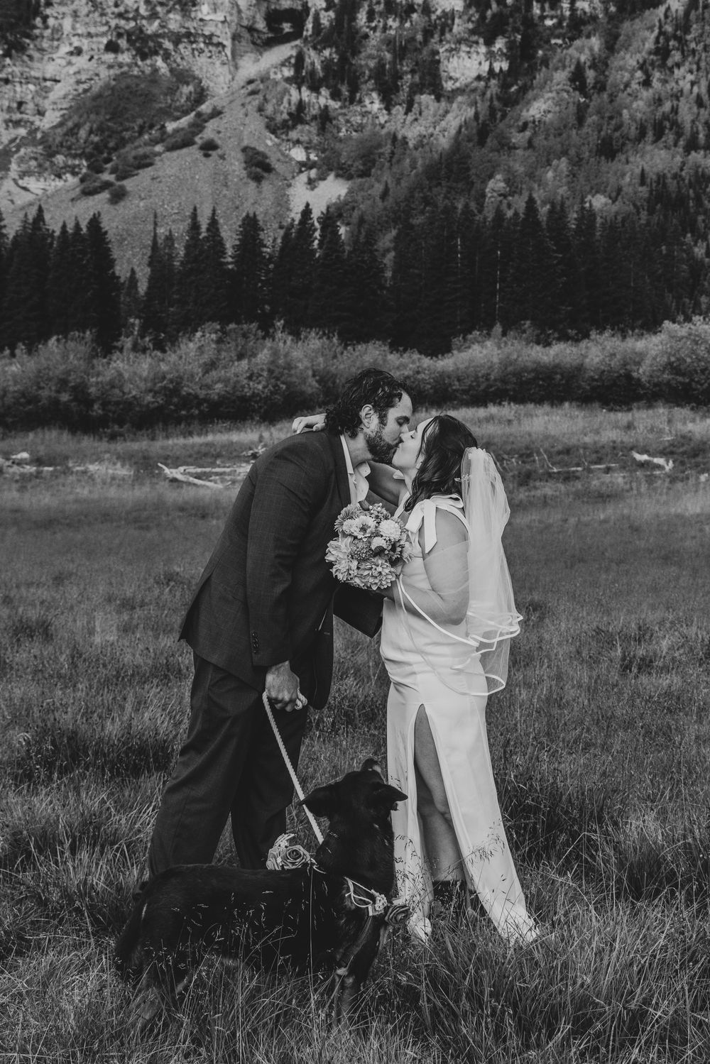 A newlywed couple shares playful moments in a mountain meadow with evergreen trees and rolling hills in the background.