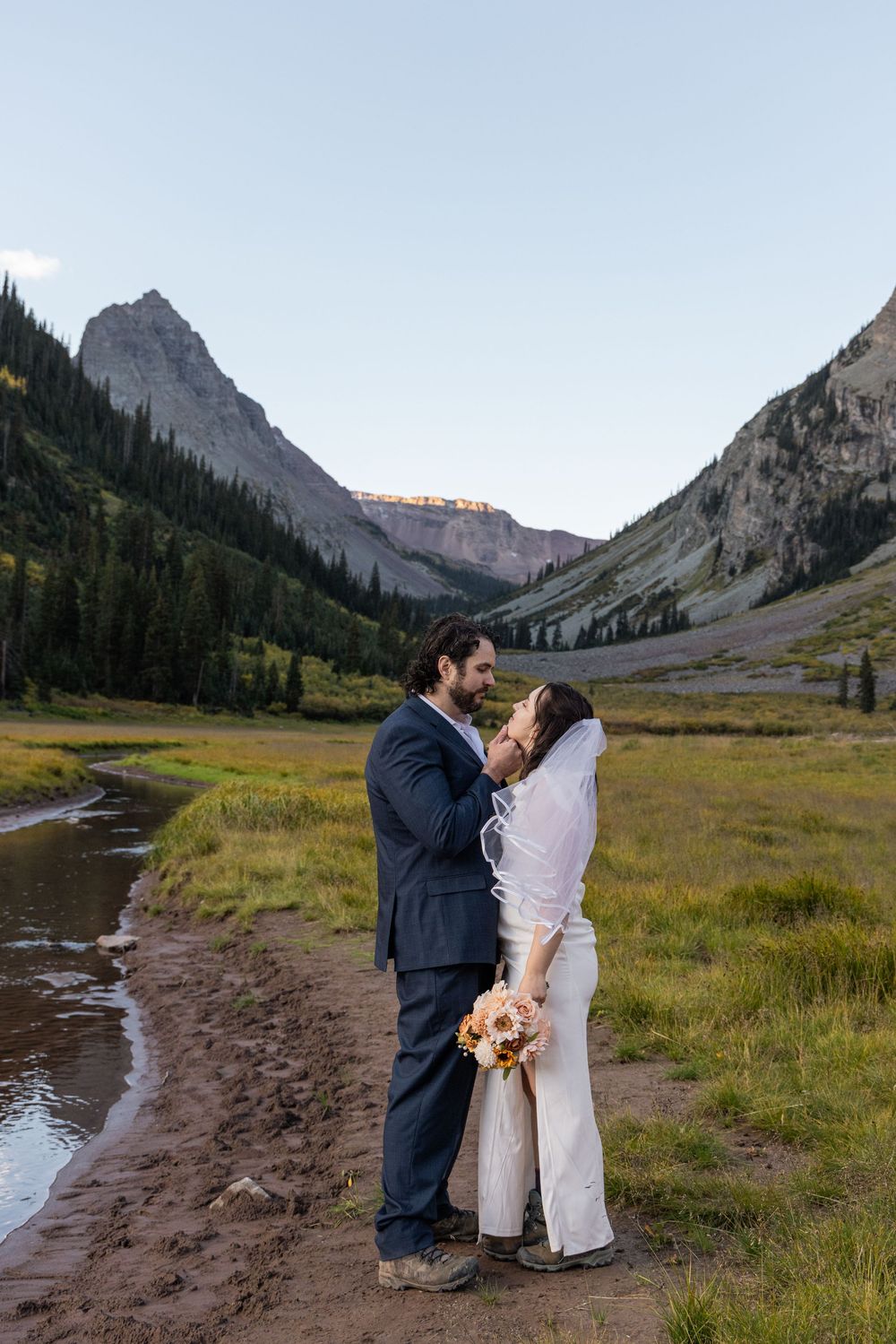 Two people share an intimate moment in formal attire against a majestic mountain landscape with green meadows and peaks.