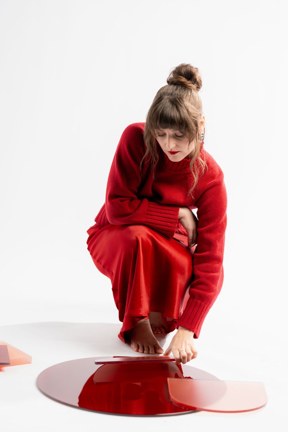 Person in an oversized red sweater dress crouches near a reflective red puddle against white background.
