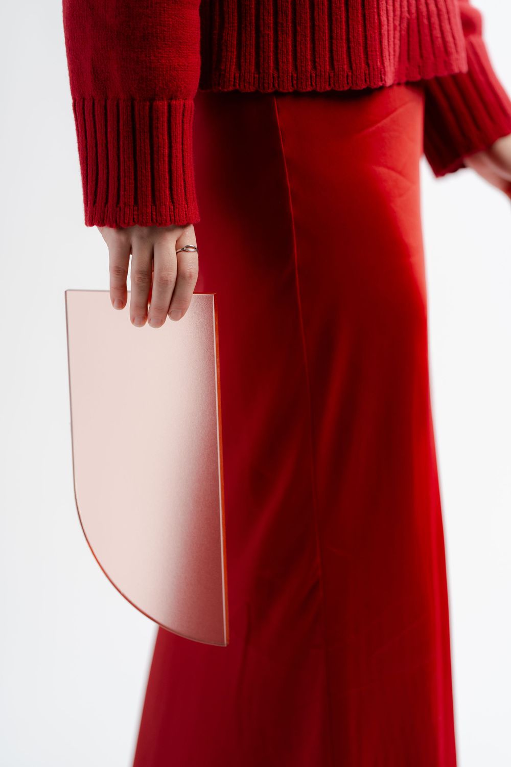 Person in red sweater and matching pants holds a curved blush pink handbag against white background.