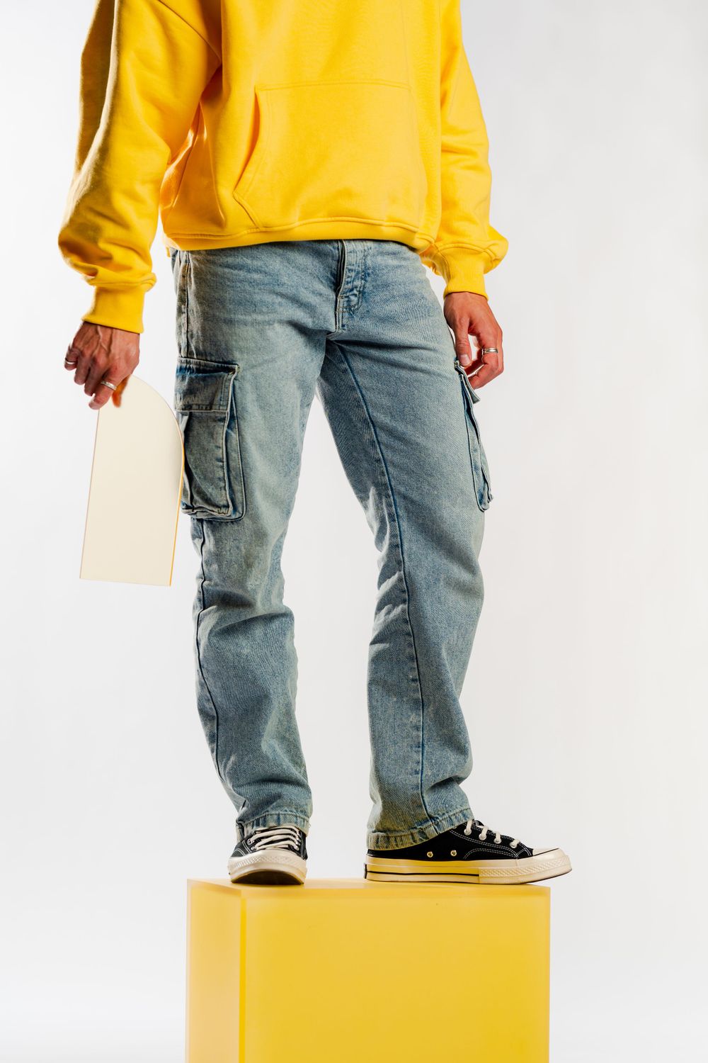 Person in yellow hoodie and cargo jeans stands on yellow cube wearing black sneakers against white background.