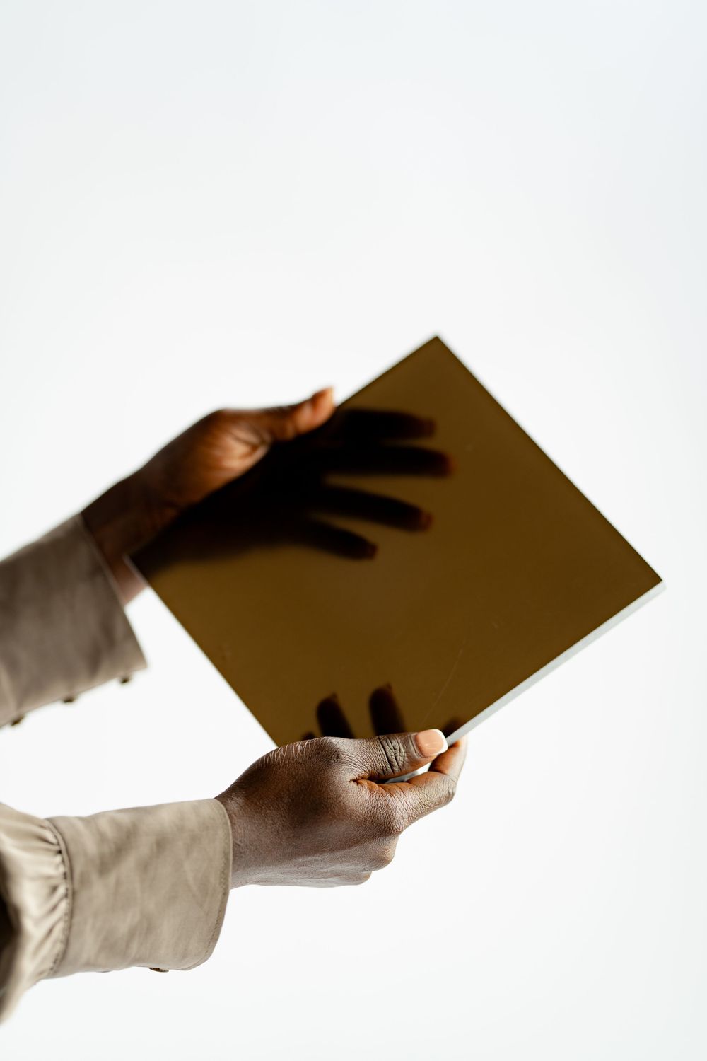 Hands holding a reflective gold-tinted mirror or glass panel against a white background showing a shadow.