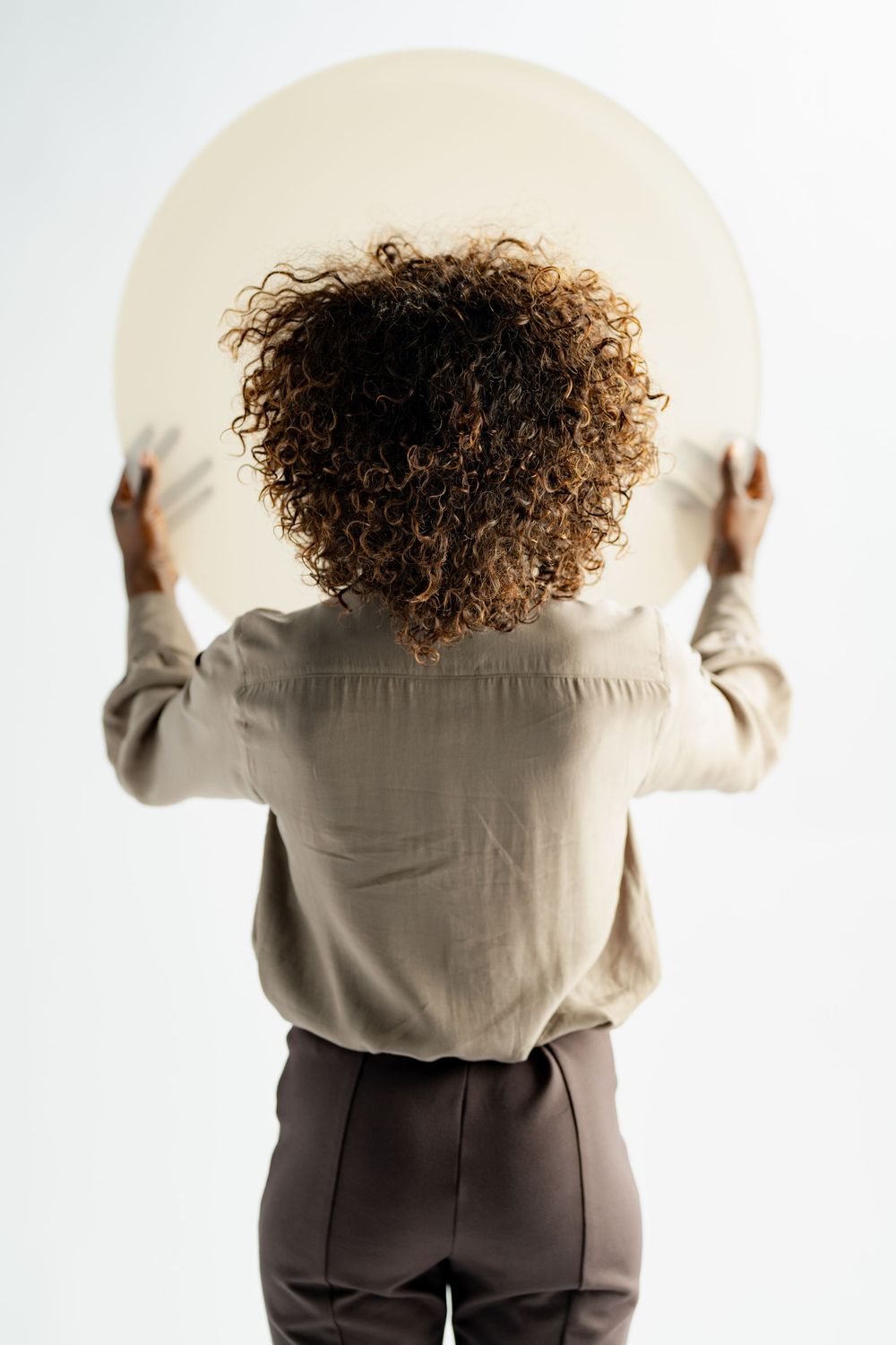 Back view of a person with curly hair holding a large white circular object against a light background.