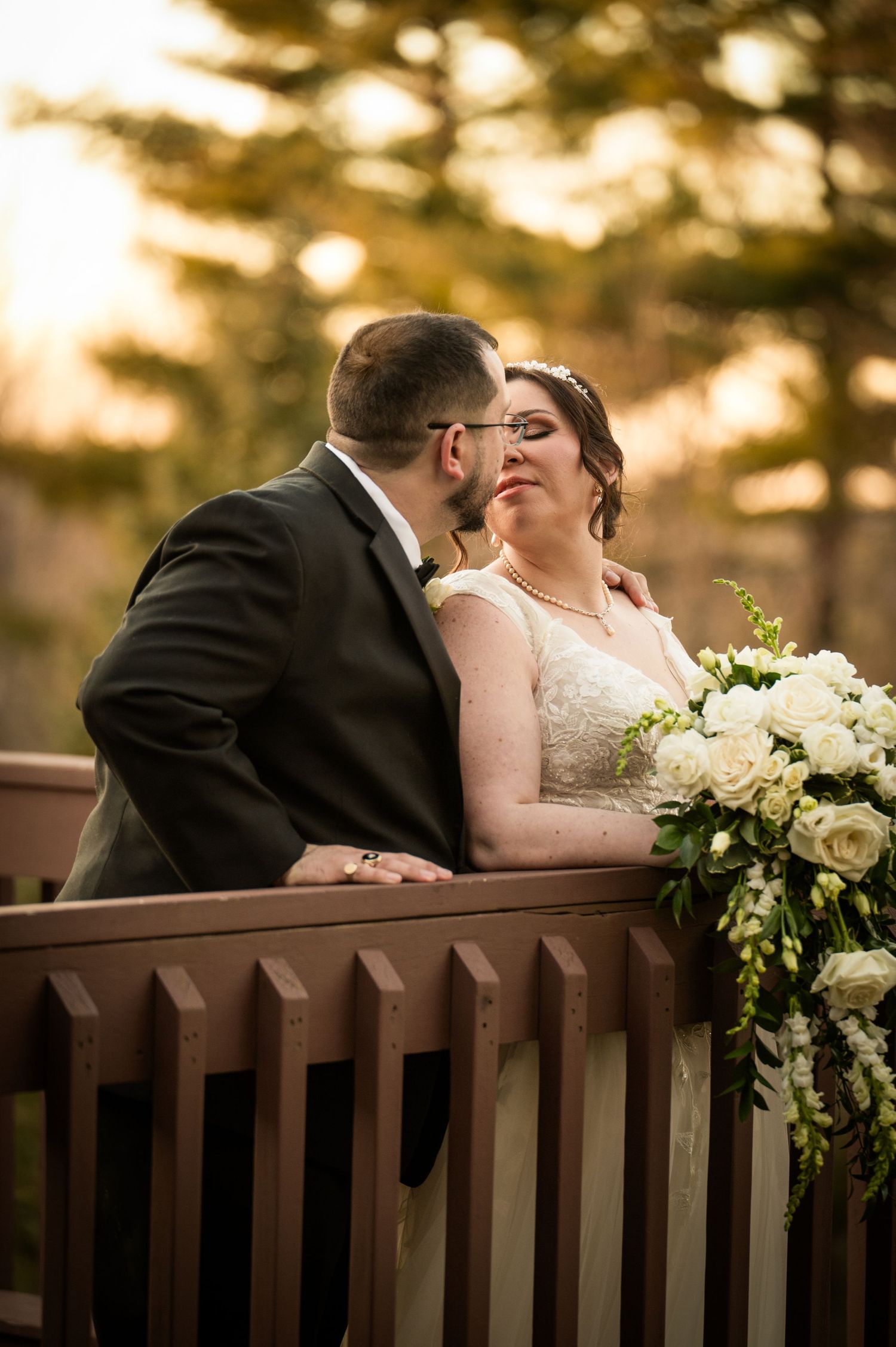 A tender wedding moment captured at sunset with a beautiful white bouquet resting on a wooden railing.