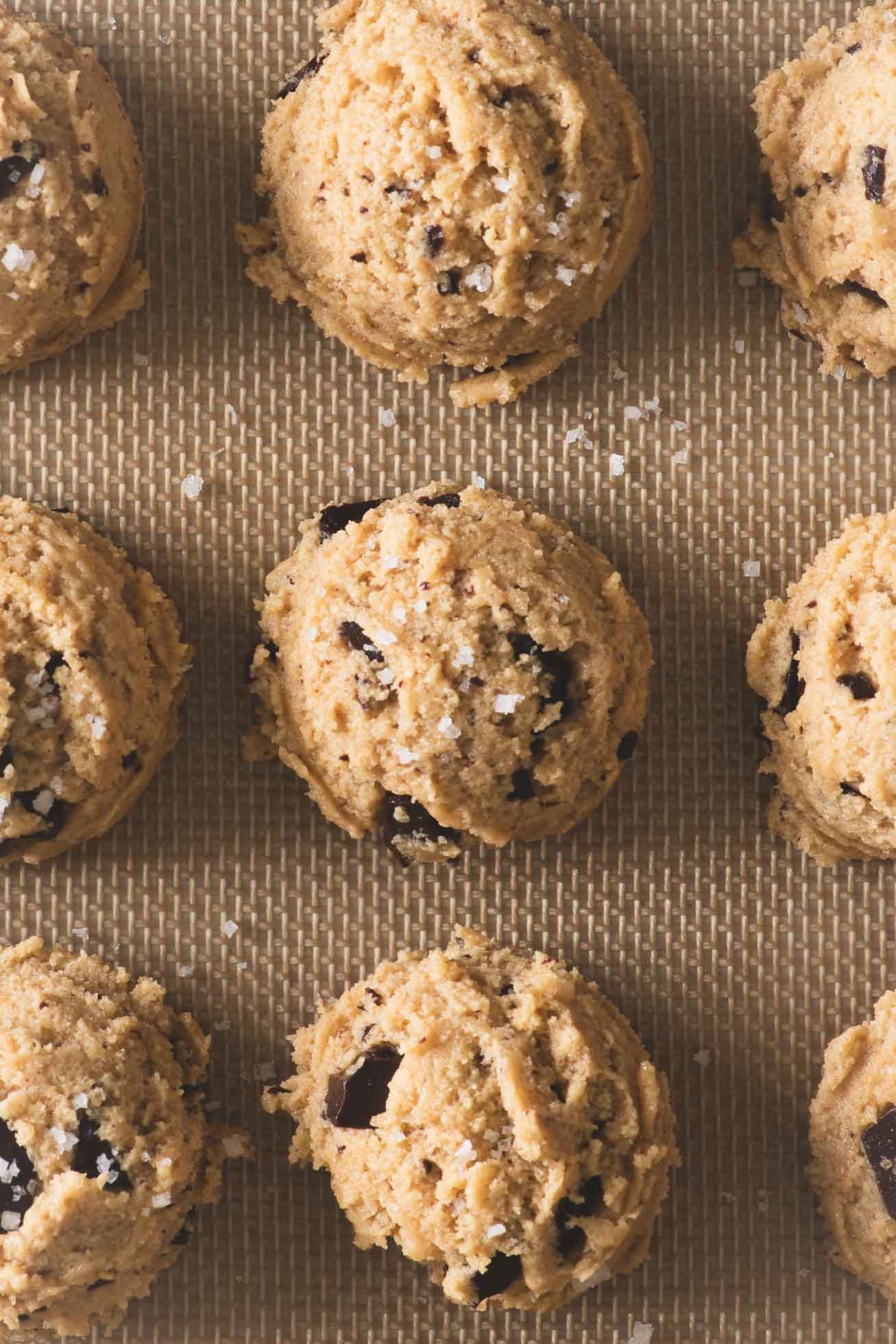 Raw chocolate chip cookie dough balls arranged on a textured baking surface ready for the oven.