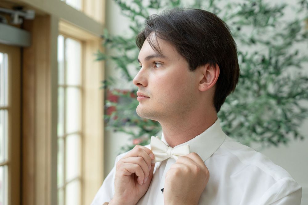 A person adjusting their bowtie while standing near a window during wedding preparations.