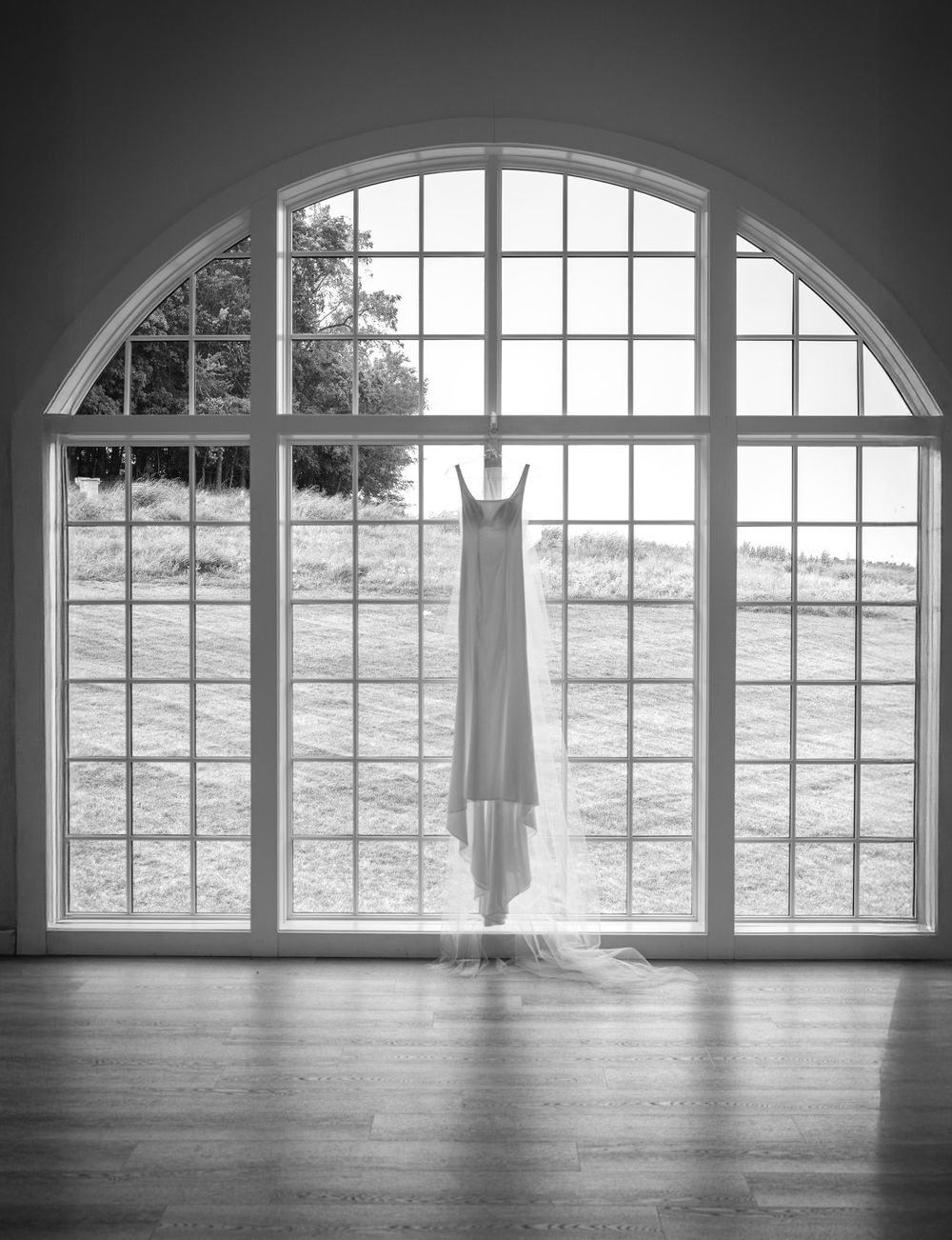 Wedding dress hangs elegantly in front of large arched window with scenic backdrop.