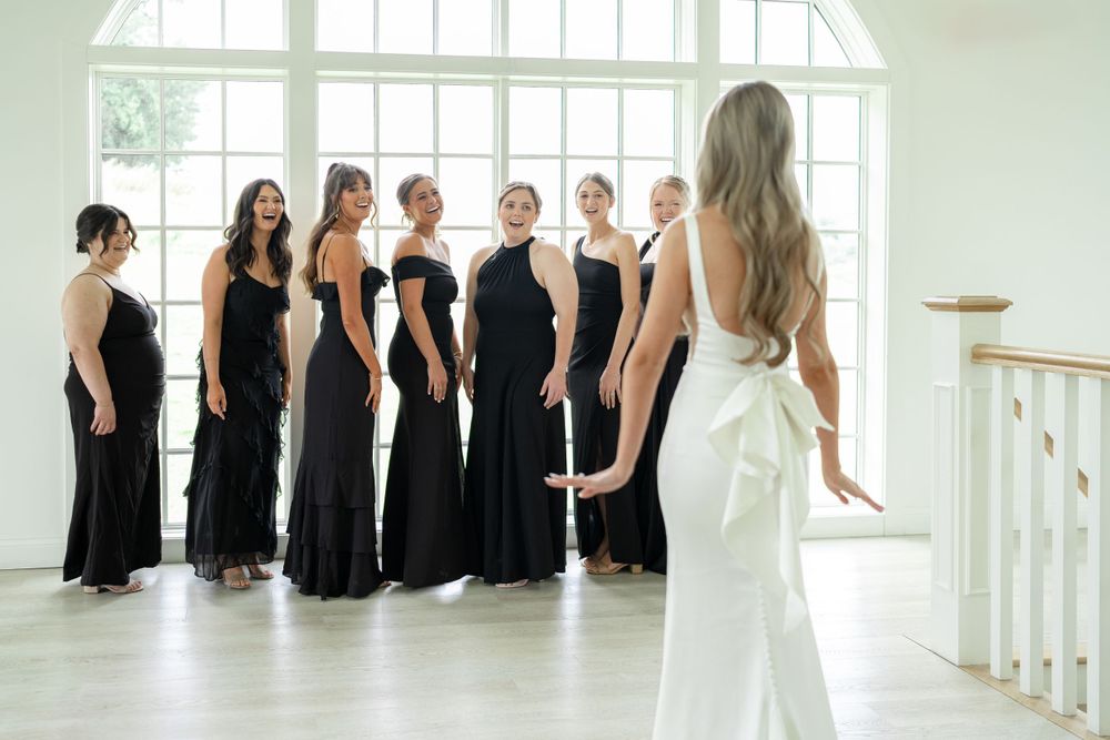 Bridesmaids in black dresses stand together while bride in white gown looks on.