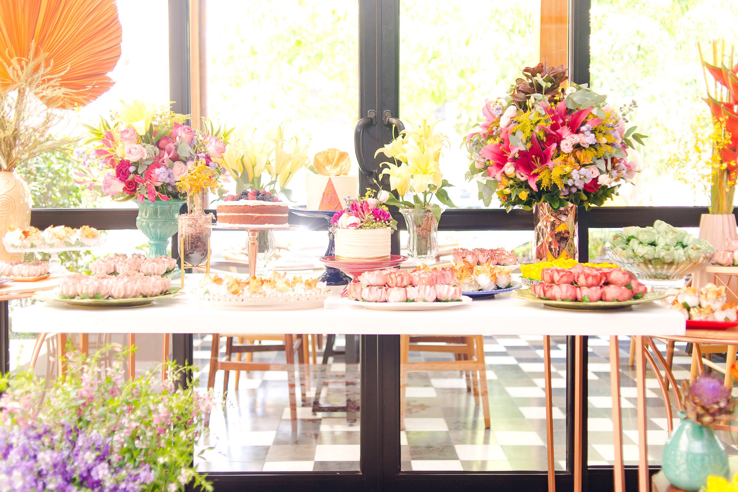 Elegant party dessert table setup with colorful floral arrangements and pastel treats on white tablecloths by sunny windows.