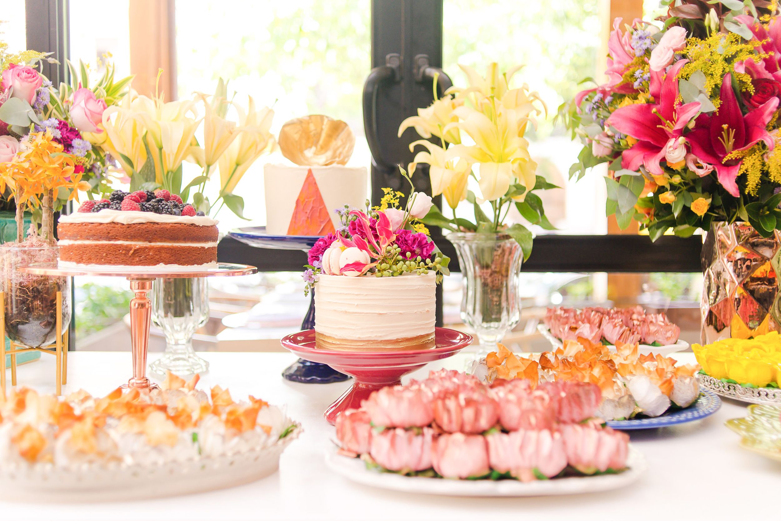 Close-up of dessert display featuring layer cakes, pink pastries and vibrant flower bouquets in soft natural lighting.