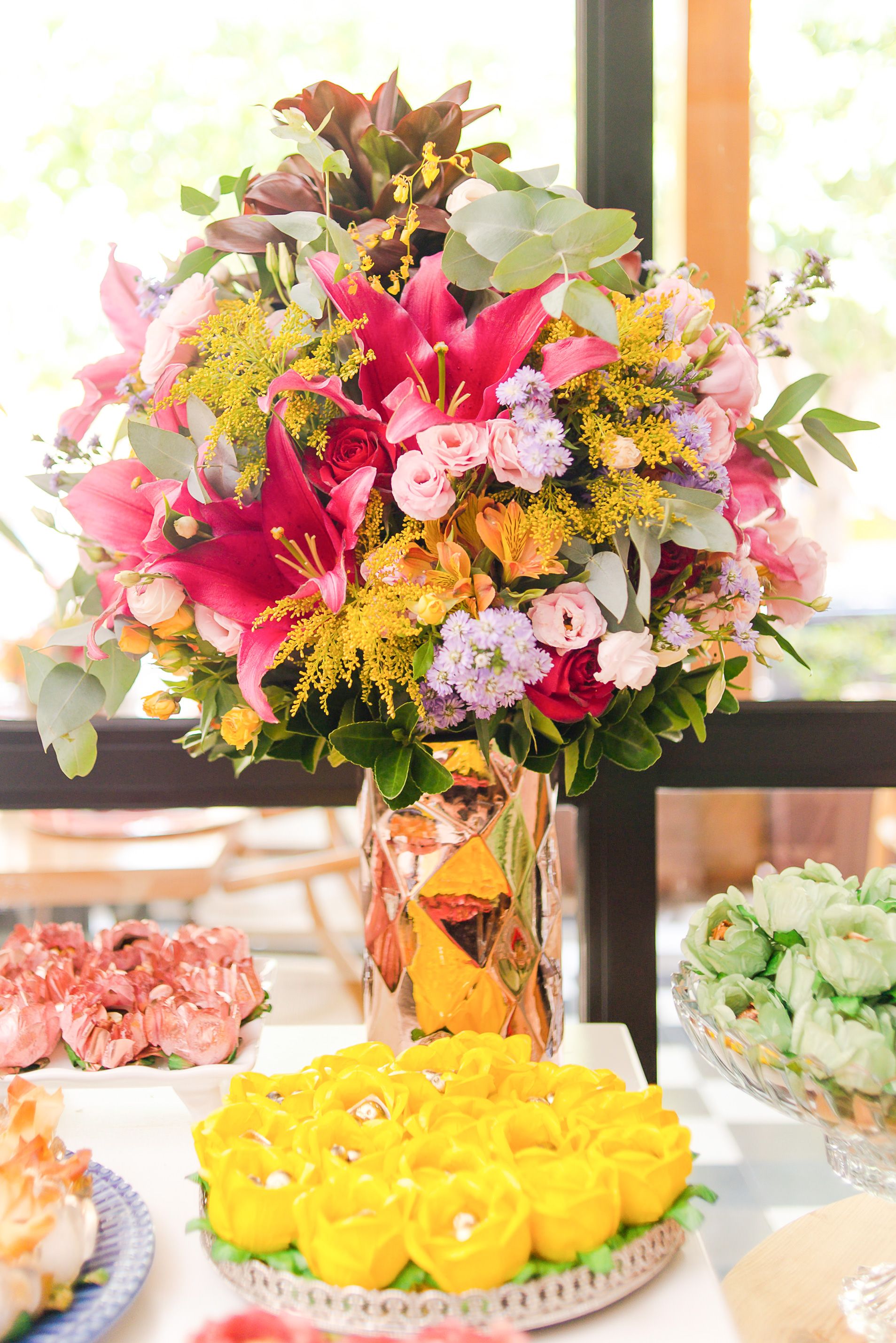 Vibrant floral centerpiece with pink and yellow blooms in geometric vase next to yellow rose desserts on display table.