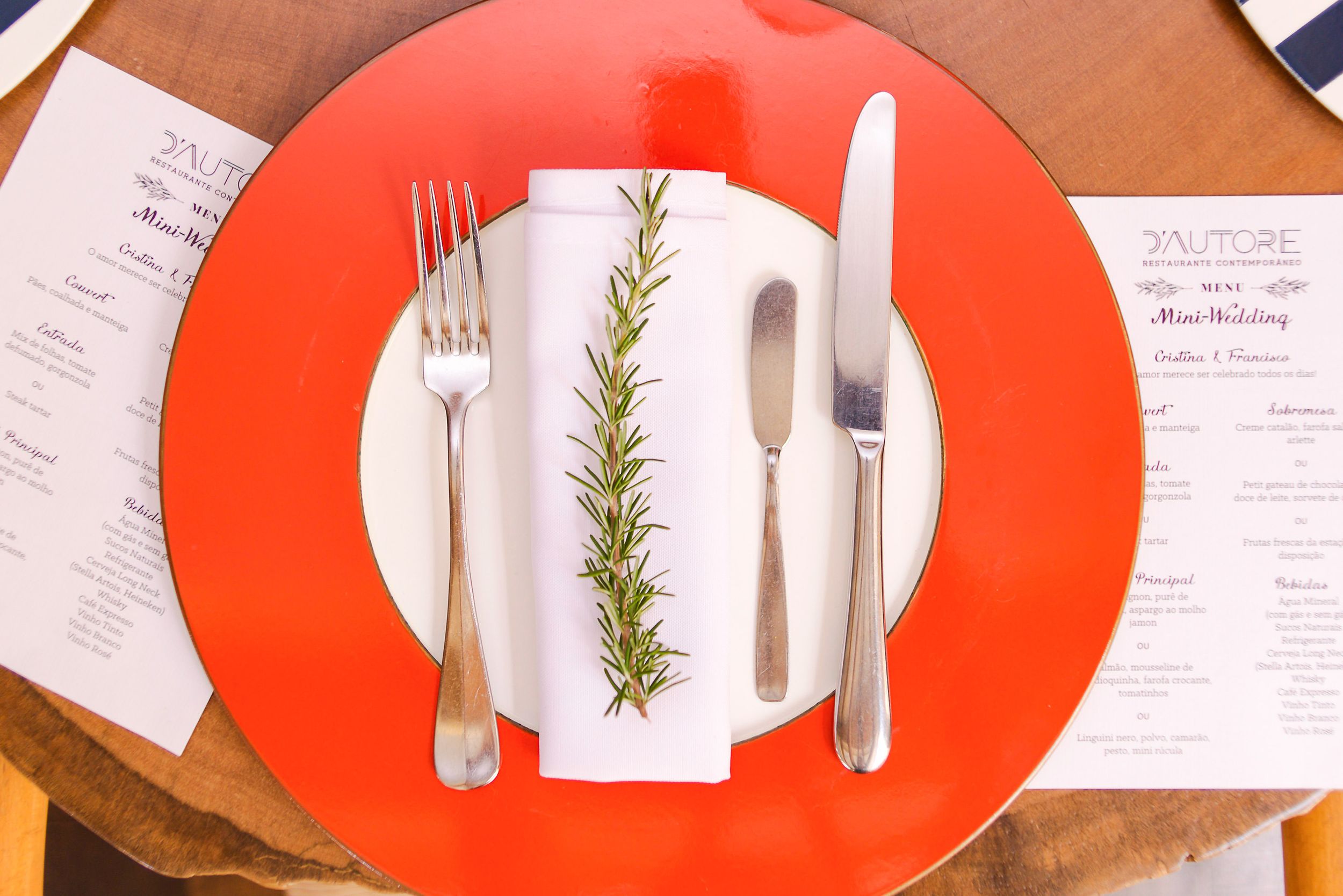 Place setting on a bright red plate with a sprig of rosemary, knife and fork on white napkin between handwritten notes.