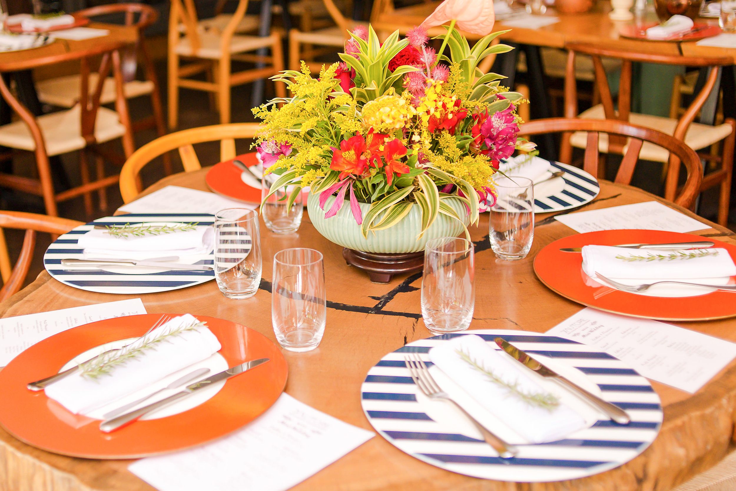 Round dining table set with orange charger plates, navy striped napkins and vibrant floral centerpiece in rustic setting.