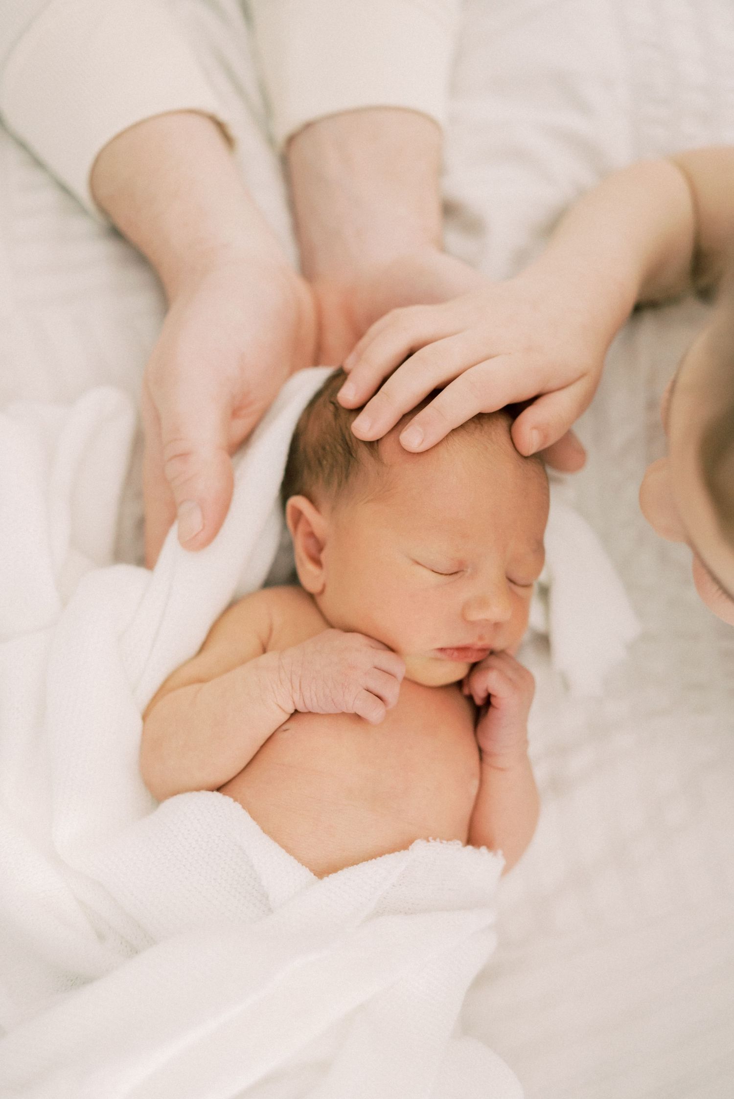 A sleeping newborn baby wrapped in soft white fabric while gentle hands tenderly touch their head.