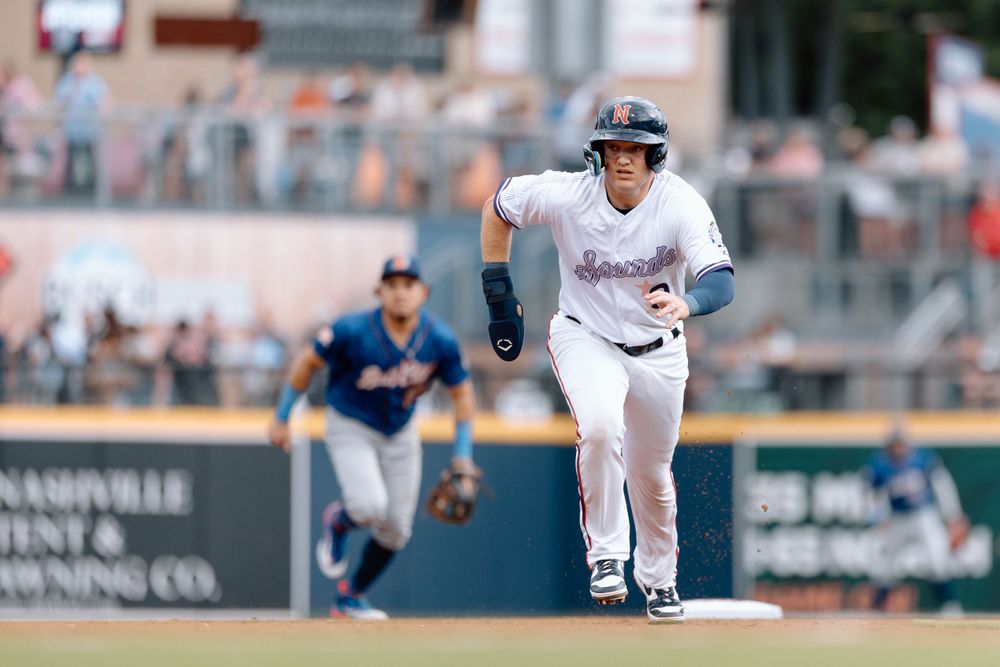 Baseball player in white uniform running between bases during a professional game with opponent in blue uniform in background.