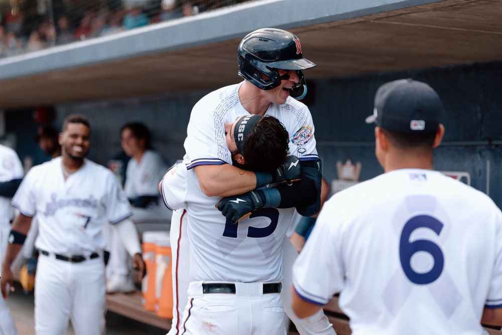 Baseball players in white uniforms interact in the dugout during a game, with one wearing a helmet and batting gear.