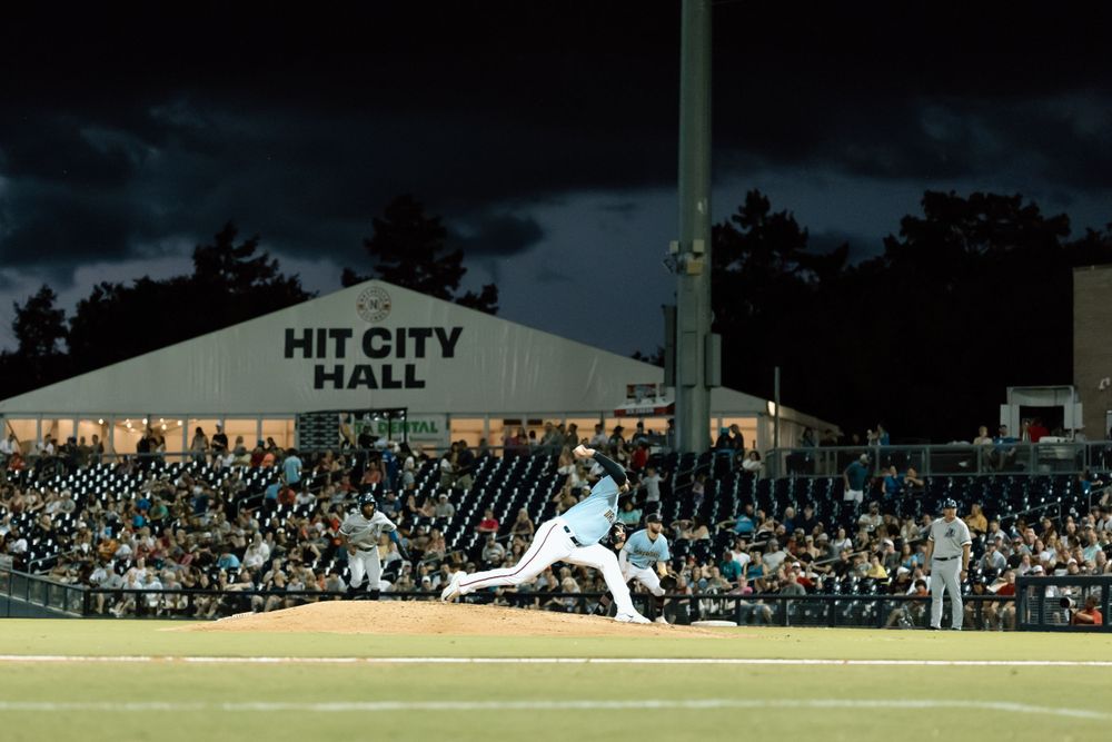 Baseball player pitches during night game at Hit City Hall stadium with illuminated stands filled with spectators.