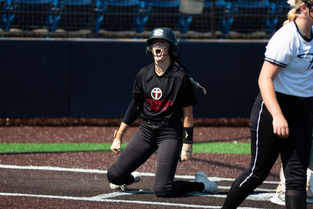 A softball player in black uniform kneels on one knee at home plate during a game at the baseball stadium.