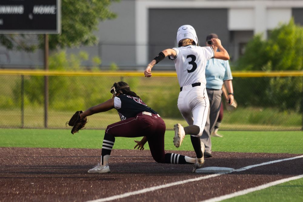Baseball player in white uniform reaches first base as baseman in maroon stretches to make the play.