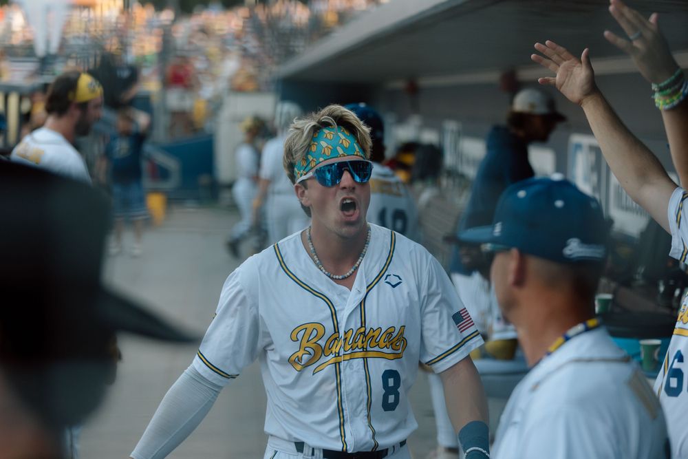 Baseball player in white jersey with reflective sunglasses celebrates in dugout with teammates at stadium.