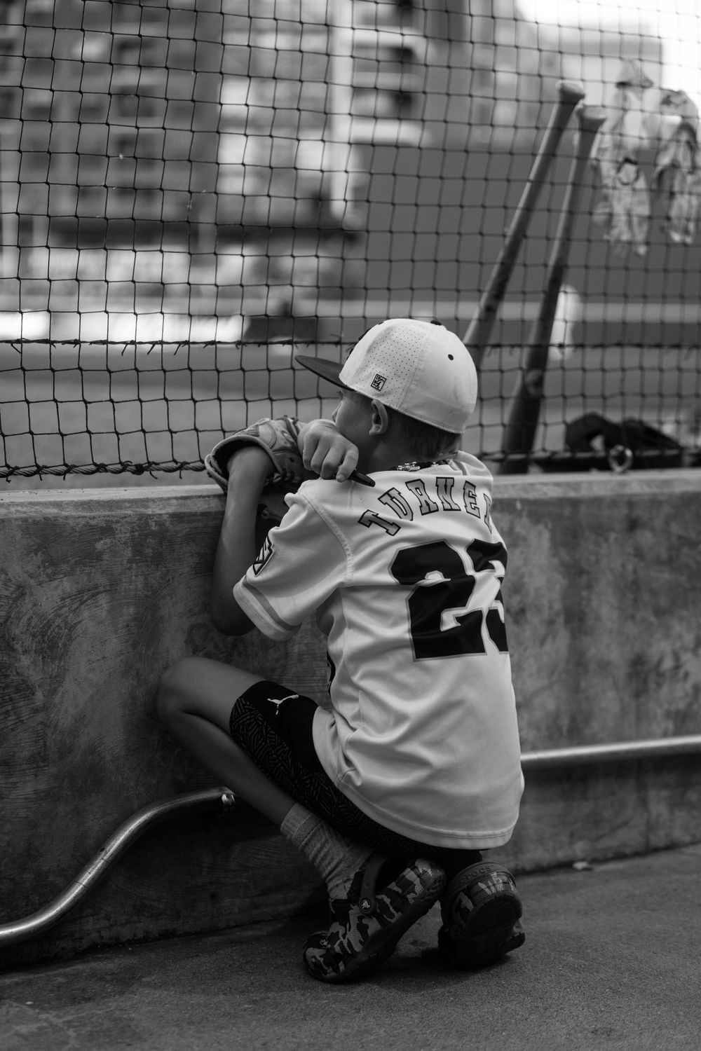 A young baseball player wearing jersey number 2 crouches by a batting cage net in black and white photography.