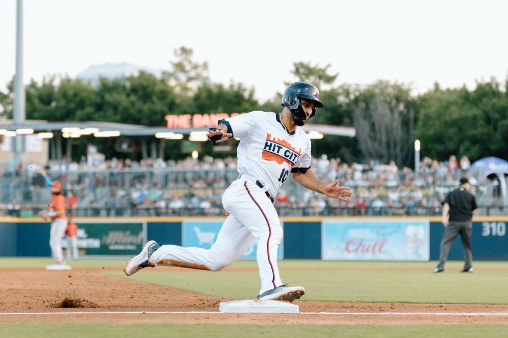 Baseball player in white uniform swings bat during game at outdoor stadium on sunny day.