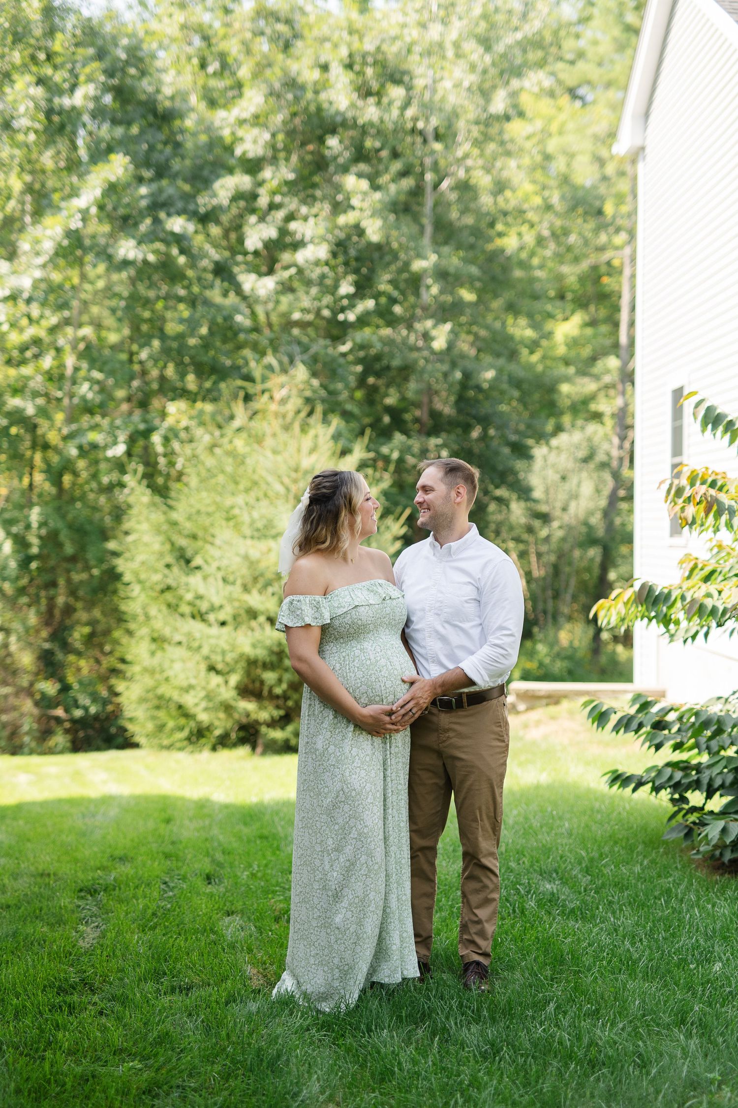Expecting couple poses outdoors on a lush lawn beside a white house on a sunny summer day.