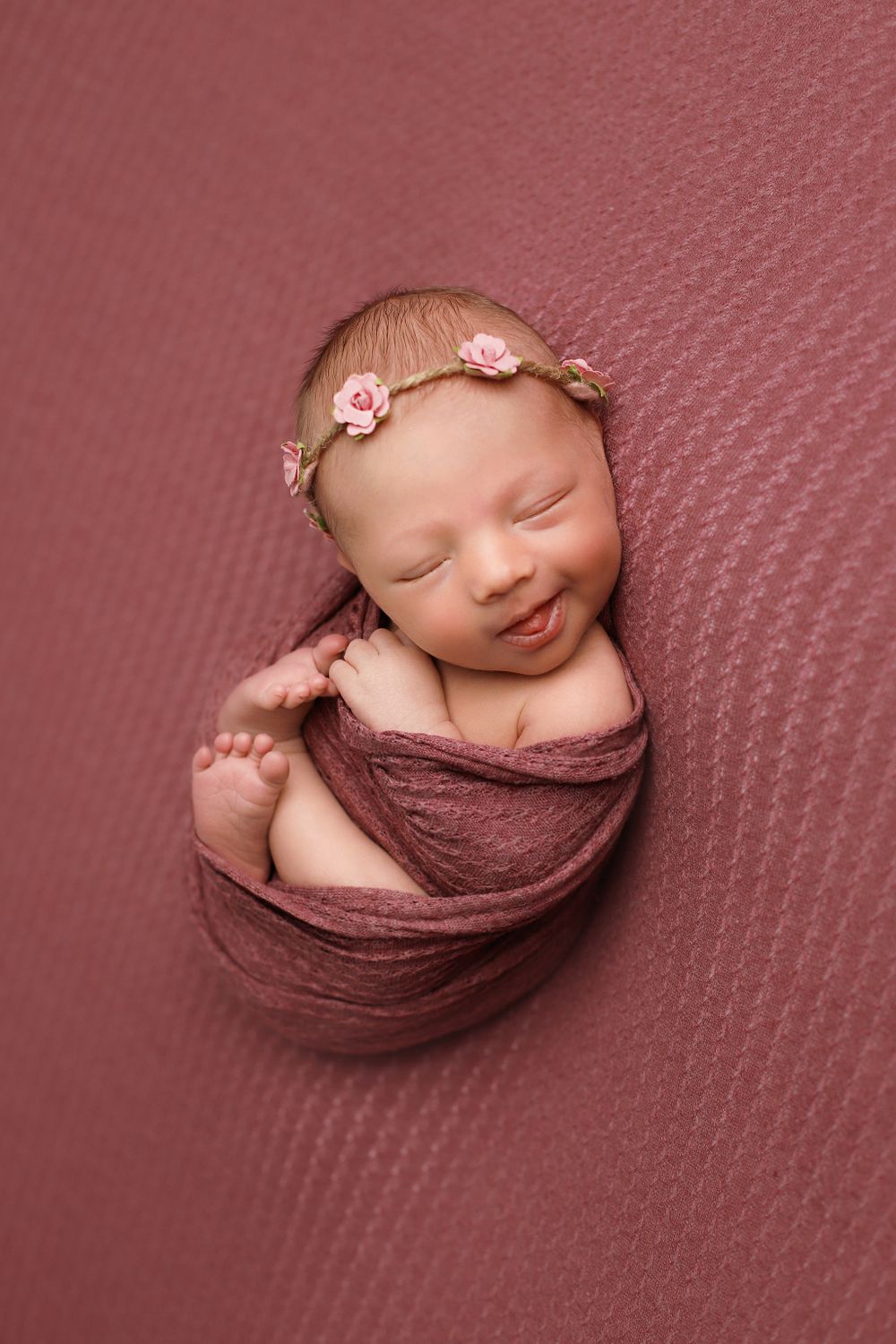 Newborn baby swaddled in mauve fabric wearing delicate pink floral headband sleeps peacefully on textured backdrop.
