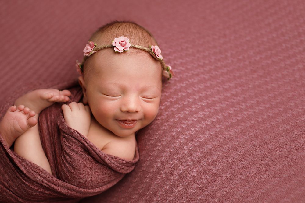 Newborn baby swaddled in brown fabric sleeping peacefully with tiny floral headband on pink textured backdrop.
