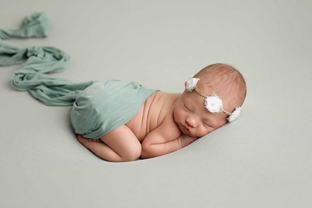 Sleeping newborn baby wearing a mint wrap and white floral headband rests curled up on neutral backdrop during photo session.