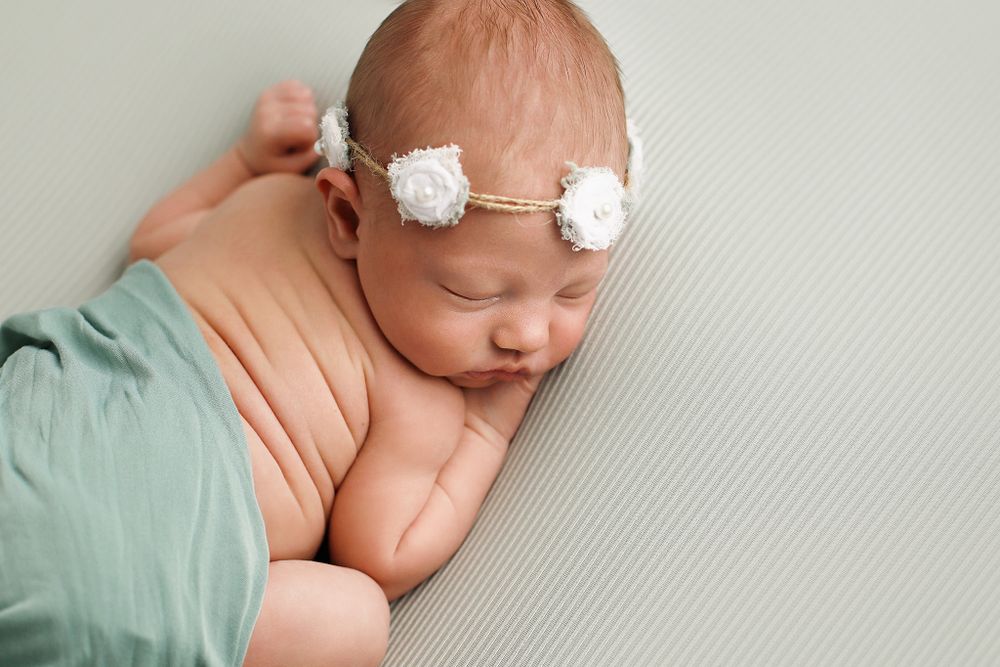 Sleeping newborn wearing delicate white floral headband wrapped in mint green fabric on textured gray background.